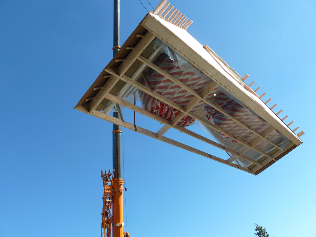 Orange kran lyfter en färdigbyggd takmodul med isolering mot en klarblå himmel.
