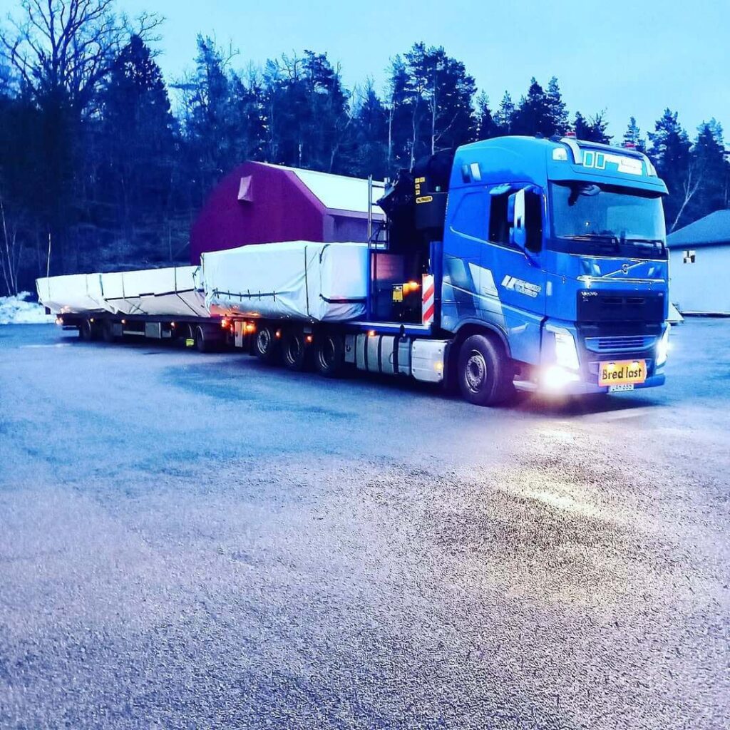 Blå Volvolastbil med skylt om bred last lastar stora takmoduler vid skymning.