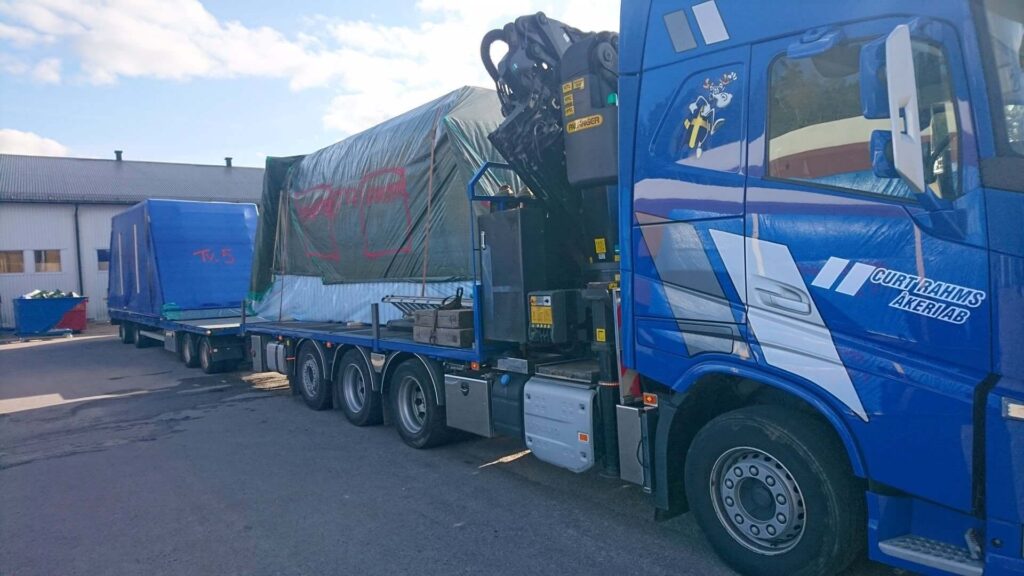 Blå lastbil med kran transporterar täckta takmoduler på släp.