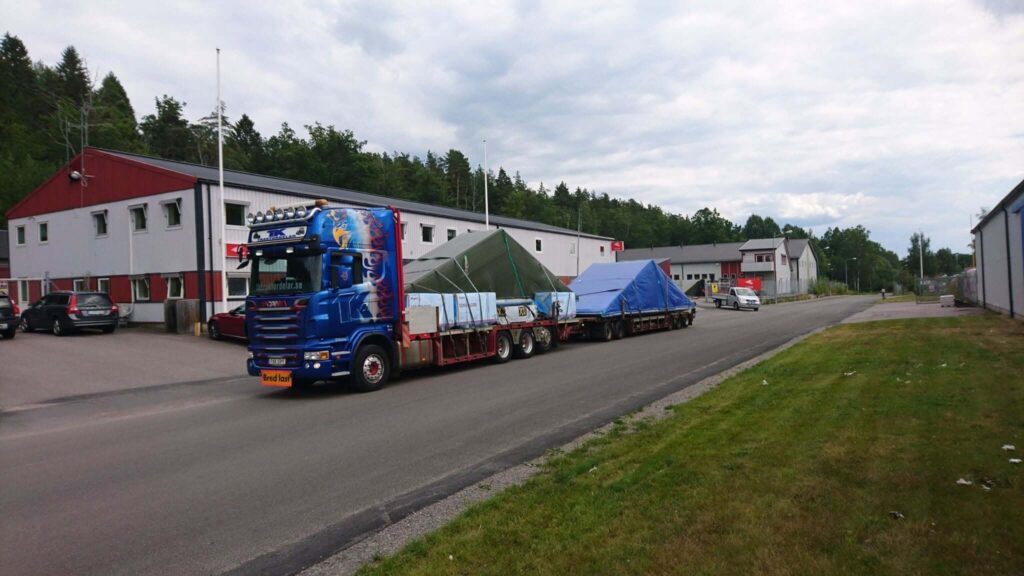 Blå Scania-lastbil med skylt om bred last transporterar täckta takmoduler.